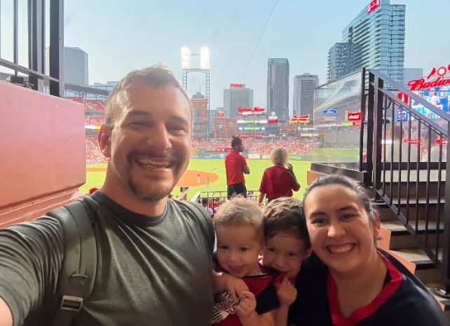 Ian at a Cardinals game with his two boys and his wife, the ballpark and the St. Louis skyline behind them